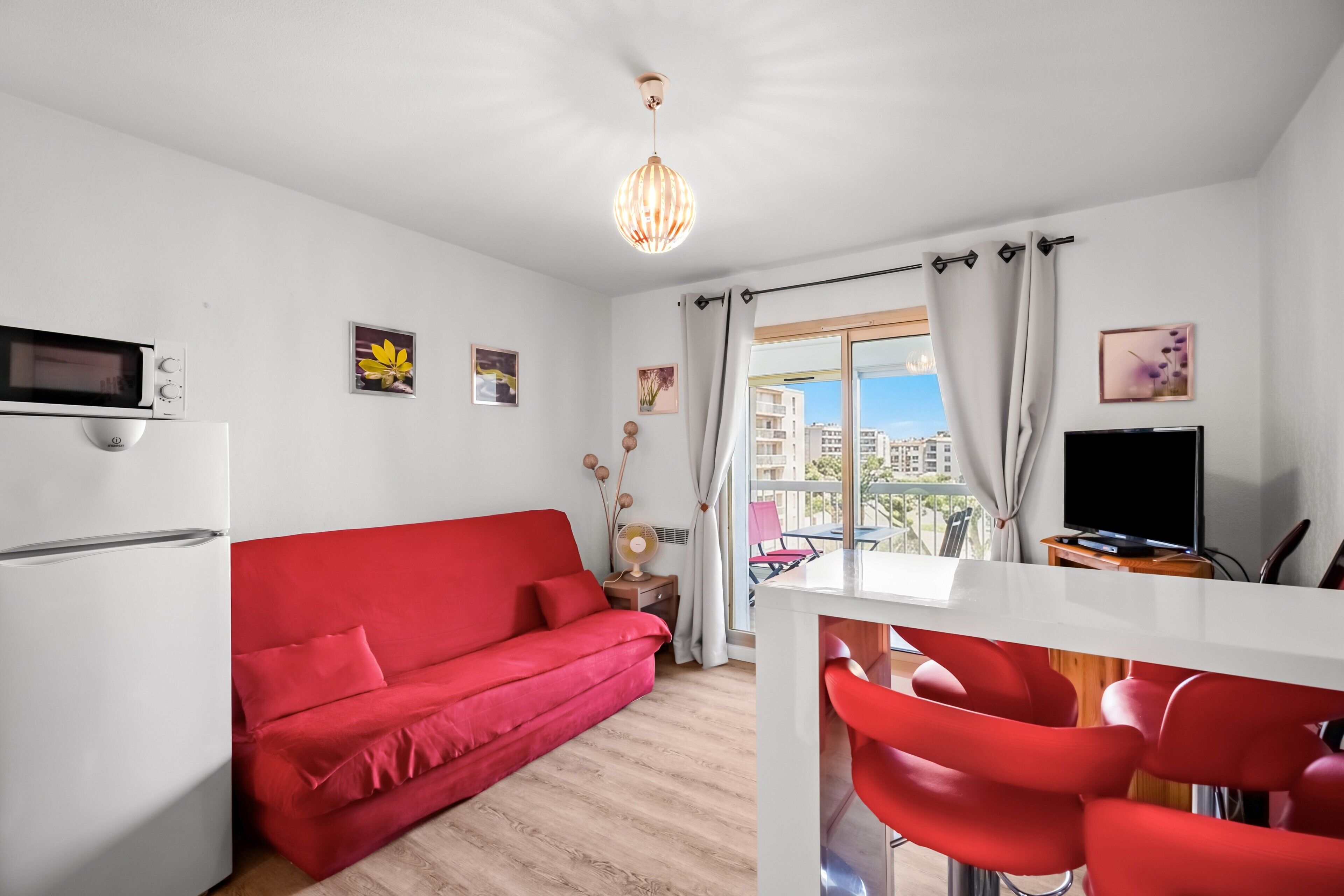 A modern living room with a red sofa, a white dining table with red chairs, a flat-screen TV, and a view of buildings through large windows.