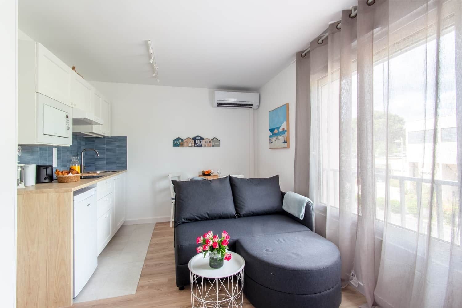 Ein zeitgemäßer Wohnraum mit Küchenzeile, einer Couch, einem kleinen Tisch mit Blumen und einem großen Fenster mit Vorhängen.