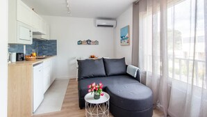 Ein zeitgemäßer Wohnraum mit Küchenzeile, einer Couch, einem kleinen Tisch mit Blumen und einem großen Fenster mit Vorhängen.