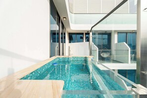 A modern indoor swimming pool with clear blue tiles, a wooden deck, and a glass balustrade. - Luxury 1BR with Private Pool & Balcony (Arjan)