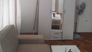 Habitación minimalista con sofá, armario, radiador, ventilador de pie y una pequeña mesa con planta en maceta.
