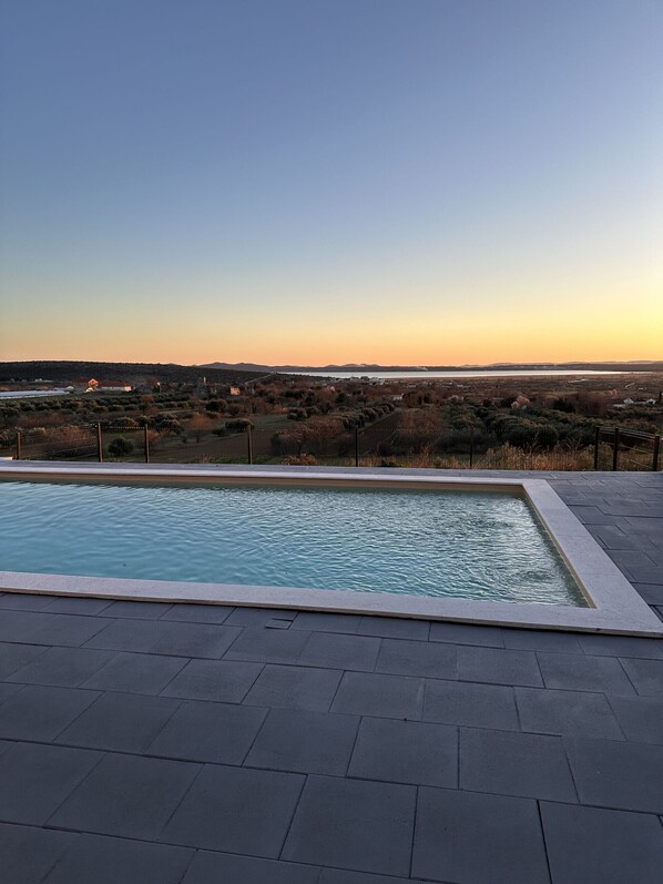 Piscina retangular com um deck ladrilhado em primeiro plano e vista para uma paisagem rural com colinas e um corpo d'água ao fundo.