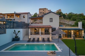 Uma vila moderna com piscina, área de estar externa e vista para ruínas históricas ao fundo.