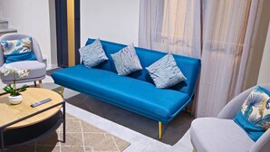 Ein modernes Wohnzimmer mit einem blauen Sofa, zwei gemusterten Kissen, einem grauen Sessel, einem kleinen runden Tisch mit einer Fernbedienung und einer Topfpflanze.