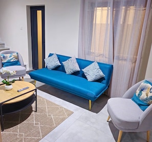 A modern living room with a blue sofa, two patterned pillows, a grey armchair, a small round table with a remote control, and a potted plant. - Charming 3-bedroom villa in peaceful Albion with pool, Villa Serenity (Albion)