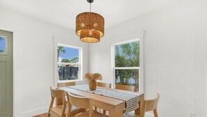 A dining area with a wooden table and chairs, a woven pendant light, and two windows with views of palm trees and buildings.
