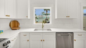 A modern kitchen with white cabinetry, a stainless steel sink, and a window with a view of a house and palm trees.