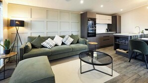 Een moderne woonkamer met een groene bank, een glazen salontafel en een keuken met donkere kastjes en een inbouwoven.