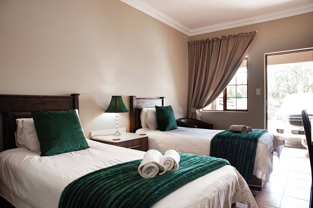 Une chambre d’hôtel avec deux lits, chacun recouvert d’un plaid vert et agrémenté d’oreillers, une table de chevet avec une lampe et une fenêtre avec des rideaux.
