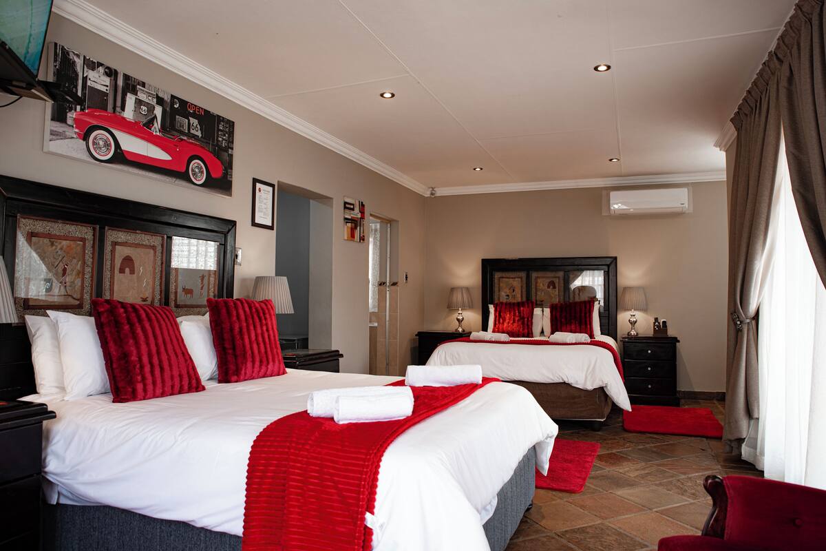 Une chambre d’hôtel avec deux lits, une literie rouge et blanche, une affiche de voiture rouge accrochée au mur et un miroir au-dessus du lit.