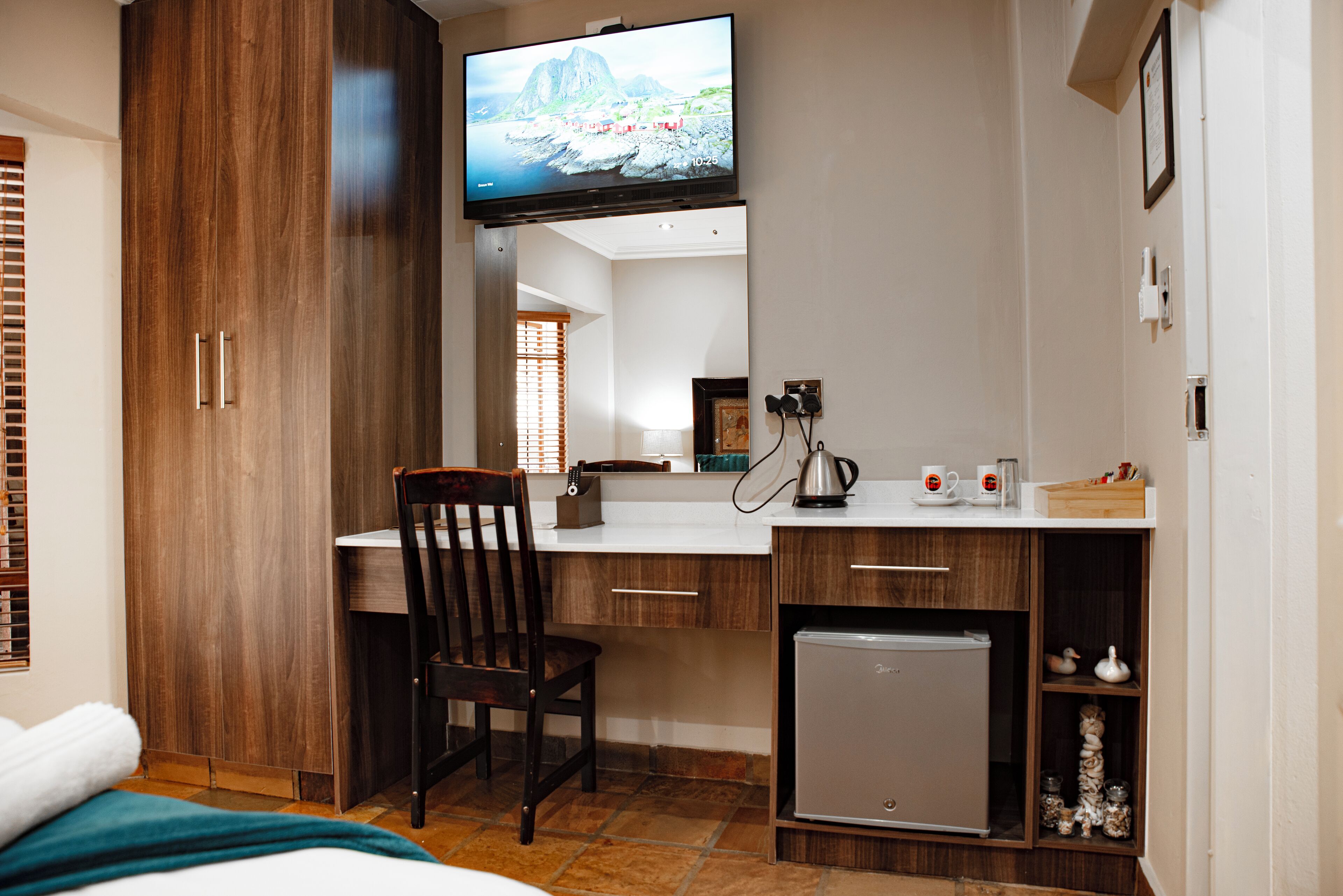 Ein kleines Hotelzimmer mit Holzschrank, einer kompakten Küchenzeile, einem Fernseher, der über der Arbeitsfläche angebracht ist, und einem einzelnen Sessel.