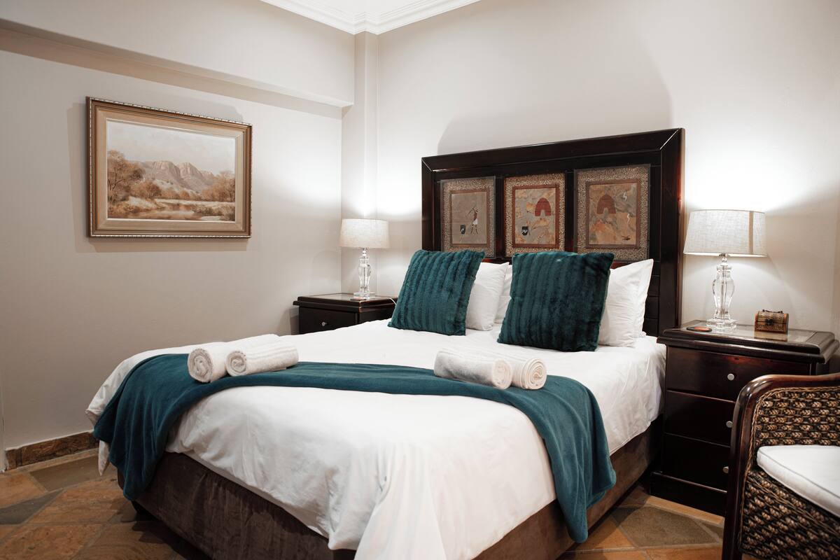 Un lit bien fait, avec du linge de lit blanc et des coussins décoratifs bleu turquoise, un tableau représentant un paysage encadré au mur et une petite table de chevet avec une lampe.