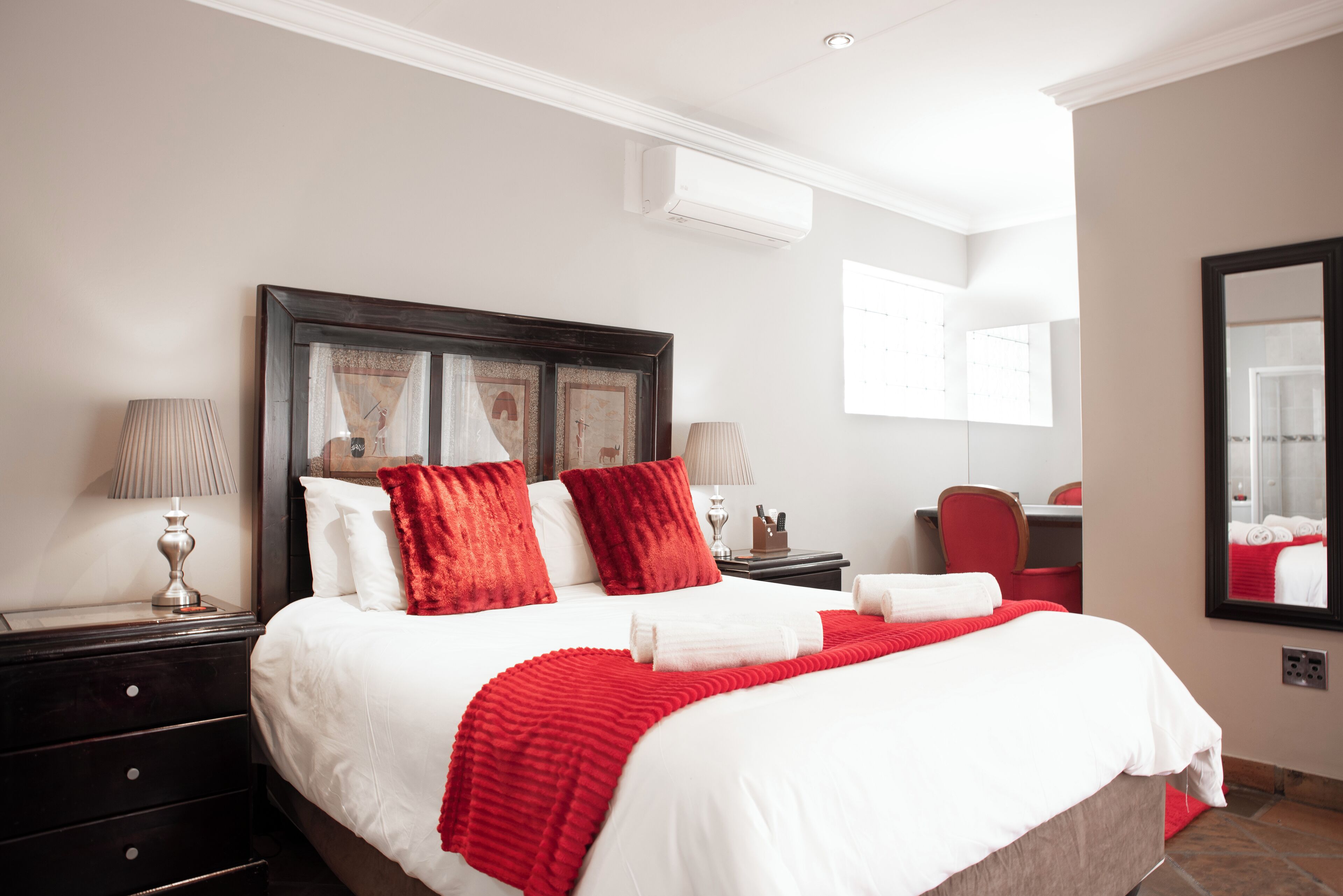 A neatly made bed with white and red bedding, a dark wooden headboard, two bedside tables with lamps, and a mirror on the wall.