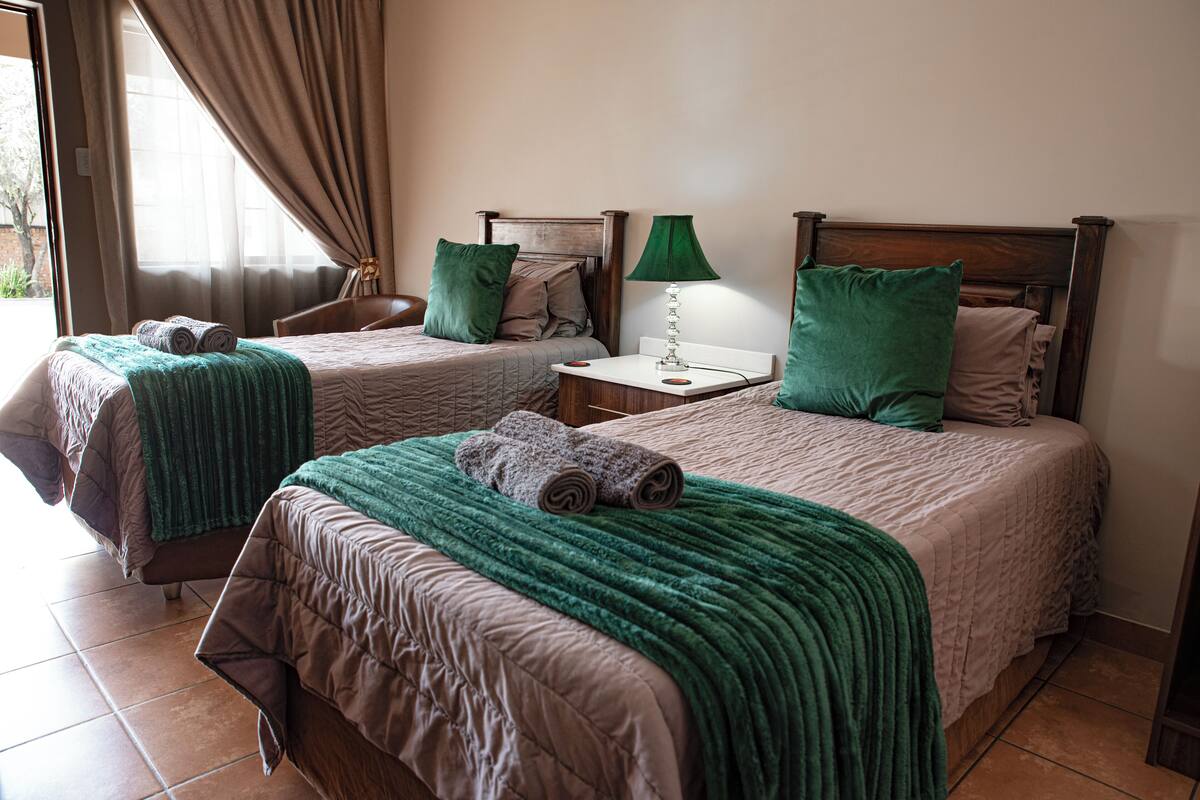 Une chambre d’hôtel avec deux lits, chacun agrémenté d’un oreiller vert et d’une serviette roulée. Une table de chevet avec une lampe se trouve entre les deux lits.