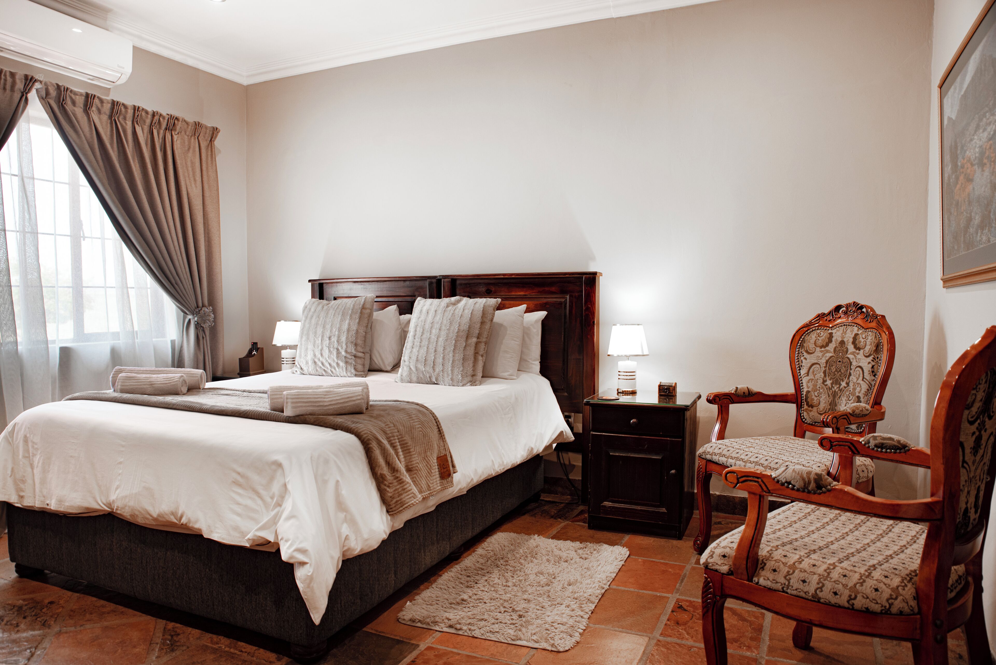 A bedroom with a large bed, a wooden nightstand, a chair, and a window with curtains.