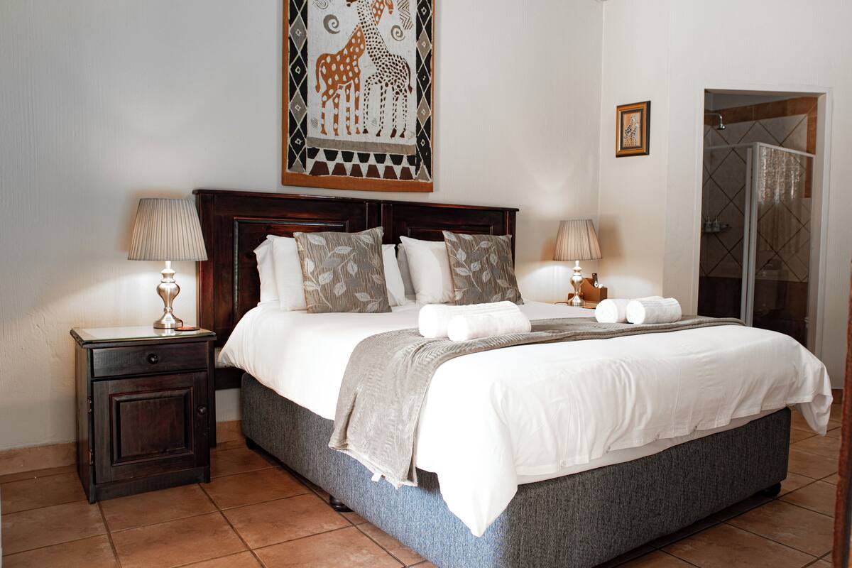 Un lit bien fait, avec du linge de lit blanc et une couverture grise, flanqué de deux tables de chevet avec des lampes, un sommier en bois et une œuvre d’art représentant des girafes accrochée au mur.