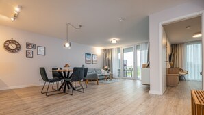 A spacious living area with a dining table, chairs, and a sofa. There are wall-mounted lights, a clock, and framed pictures.