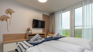 A modern hotel room with two beds, a TV, a wooden desk, and a large window offering a view of the outdoors.