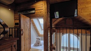 Een knus vertrek met houten wanden, dakraam, groot raam met gordijnen, en een teddybeer die bij het raam zit.