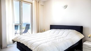 Habitación de hotel con cama, mesita de noche, lámpara y vista al mar a través de un ventanal grande.