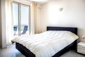Ein Hotelzimmer mit Bett, Nachttisch, Lampe und Meerblick durch ein großes Fenster.