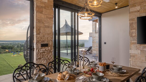 Une table à manger dressée avec des assiettes, des bols et des verres, donnant sur un paysage pittoresque et agrémentée d’une structure à toiture de chaume.