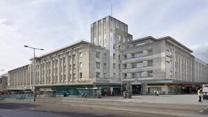 A large, multi-story building with a central tower, featuring a ground-level entrance with shops and a bus stop. - For Students Only Rooms in Plymouth (Plymouth)