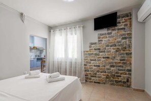Une chambre avec un lit, un mur en pierre servant d’élément décoratif, un téléviseur fixé au-dessus, et une cuisine visible à travers une porte.