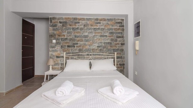 A hotel room with a stone wall, a bed with white linens, two rolled towels, a nightstand, a lamp, and a telephone. - Cozy Corner in Petralona (Athina)