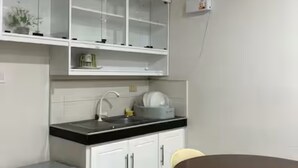 A modern kitchen with white cabinets, a black countertop, and a small refrigerator. There is a sink with a faucet, a dish rack, and a shelf above the countertop. A round table with chairs is visible in the foreground. - Relaxing Resort Stay at Ashelma Resort (Dauis)