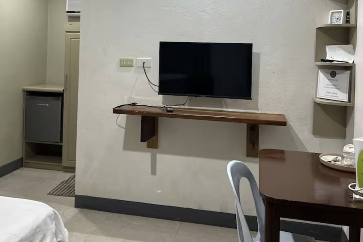 A hotel room with a flat-screen TV mounted on a wall, a small desk with a chair, and a bed with a white bedspread.