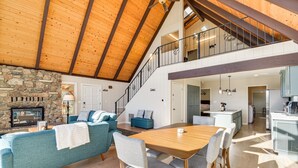 A spacious living area with a stone fireplace, a dining table with chairs, a staircase leading to an upper level, and a kitchen area.