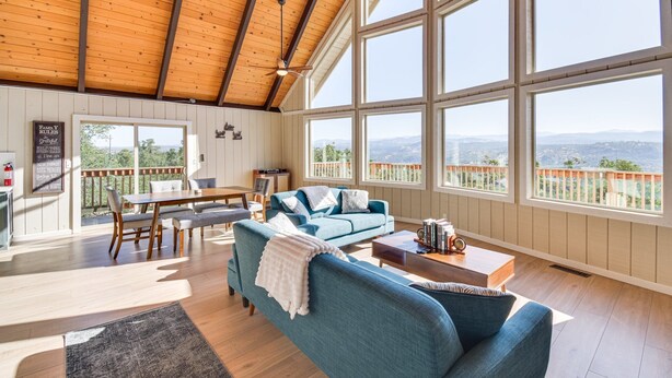 A spacious living room with a high ceiling, large windows offering a view of the mountains, a cozy blue sofa, and a dining area with a table and chairs.