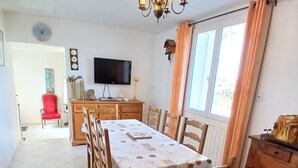 Ein Essbereich mit einem für sechs Personen gedeckten Tisch, umgeben von Holzsesseln, einer Chandelierleuchtreibe, einem an der Wand befestigten Fernseher und einem Fenster mit Vorhängen.