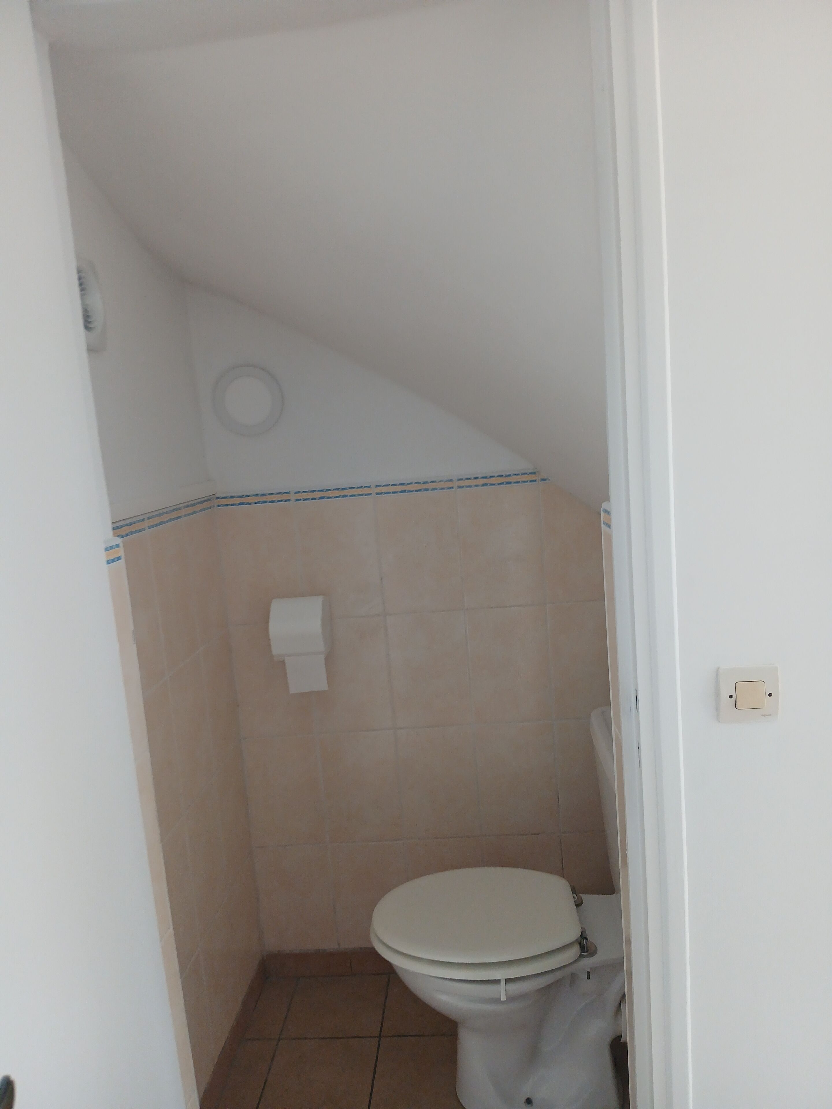 A small, narrow bathroom with a toilet, a towel dispenser, and a toilet paper holder on the wall.