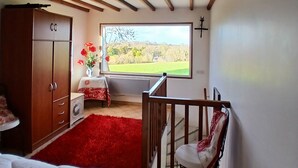 Ein Schlafzimmer mit einem Bett, einem Holzschrank, einem roten Teppich, einem kleinen Tisch mit Blumenvase und einem Fenster mit Blick auf eine grüne Landschaft.