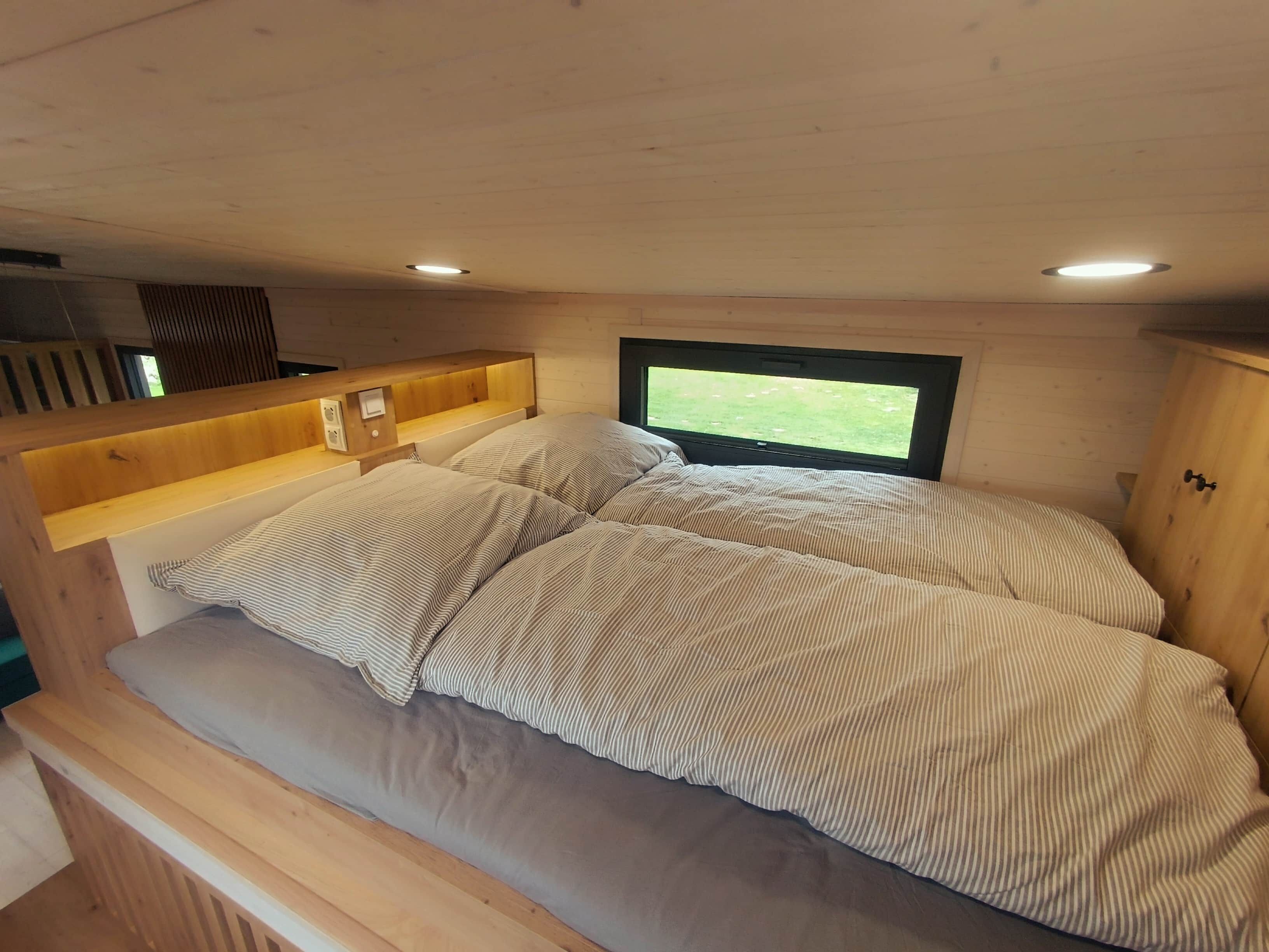 Ein Bett mit gestreifter Bettwäsche, ein Holz-Kopfteil, ein Fenster mit Blick auf Grünflächen und ein eingebautes Regal mit einer Leuchte.