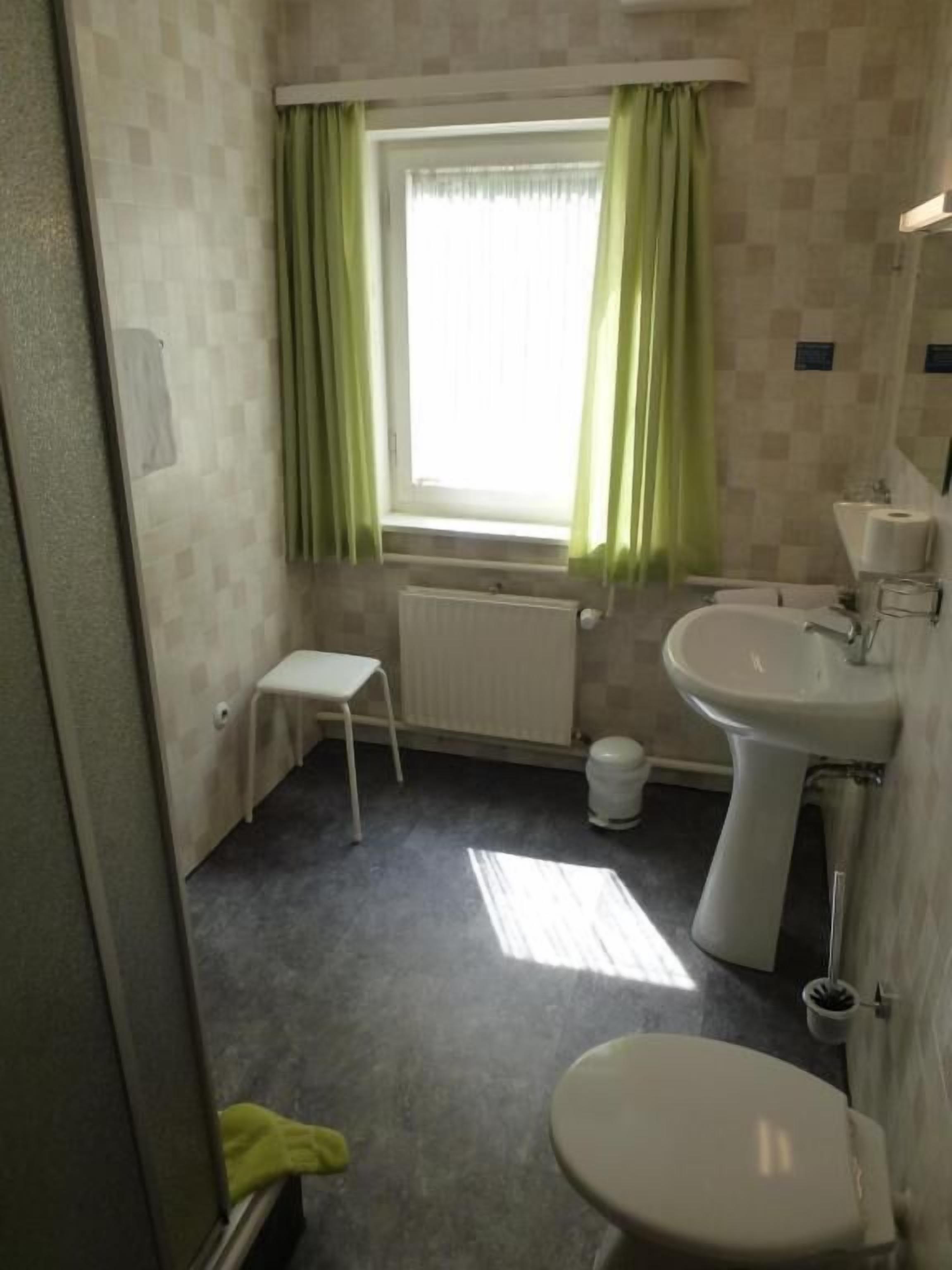 Un baño con inodoro, lavabo, ventana con cortinas, radiador y un pequeño taburete.