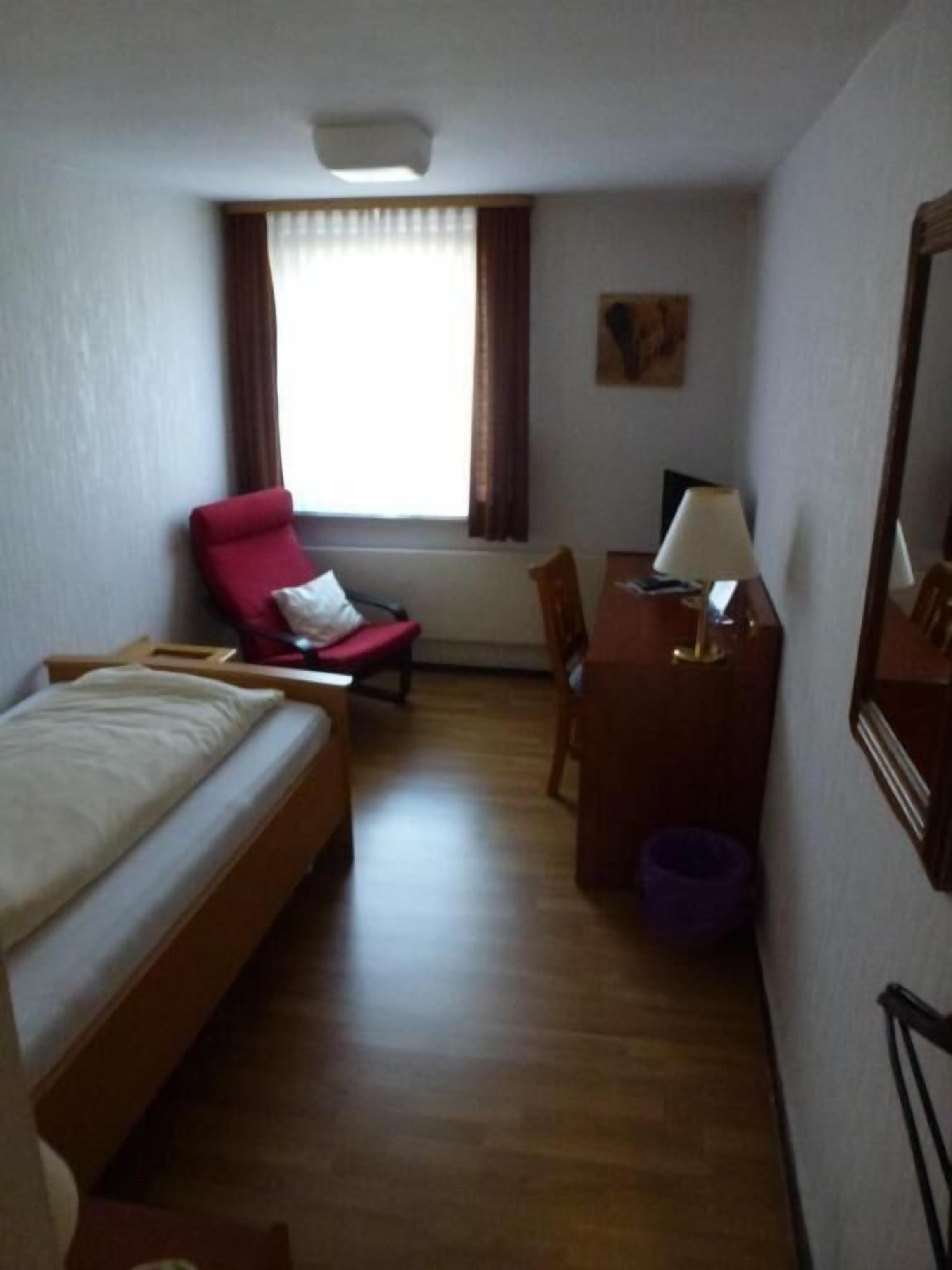 Une pièce comprenant un lit, un bureau, une chaise, une fenêtre avec des rideaux et une petite table avec une lampe.