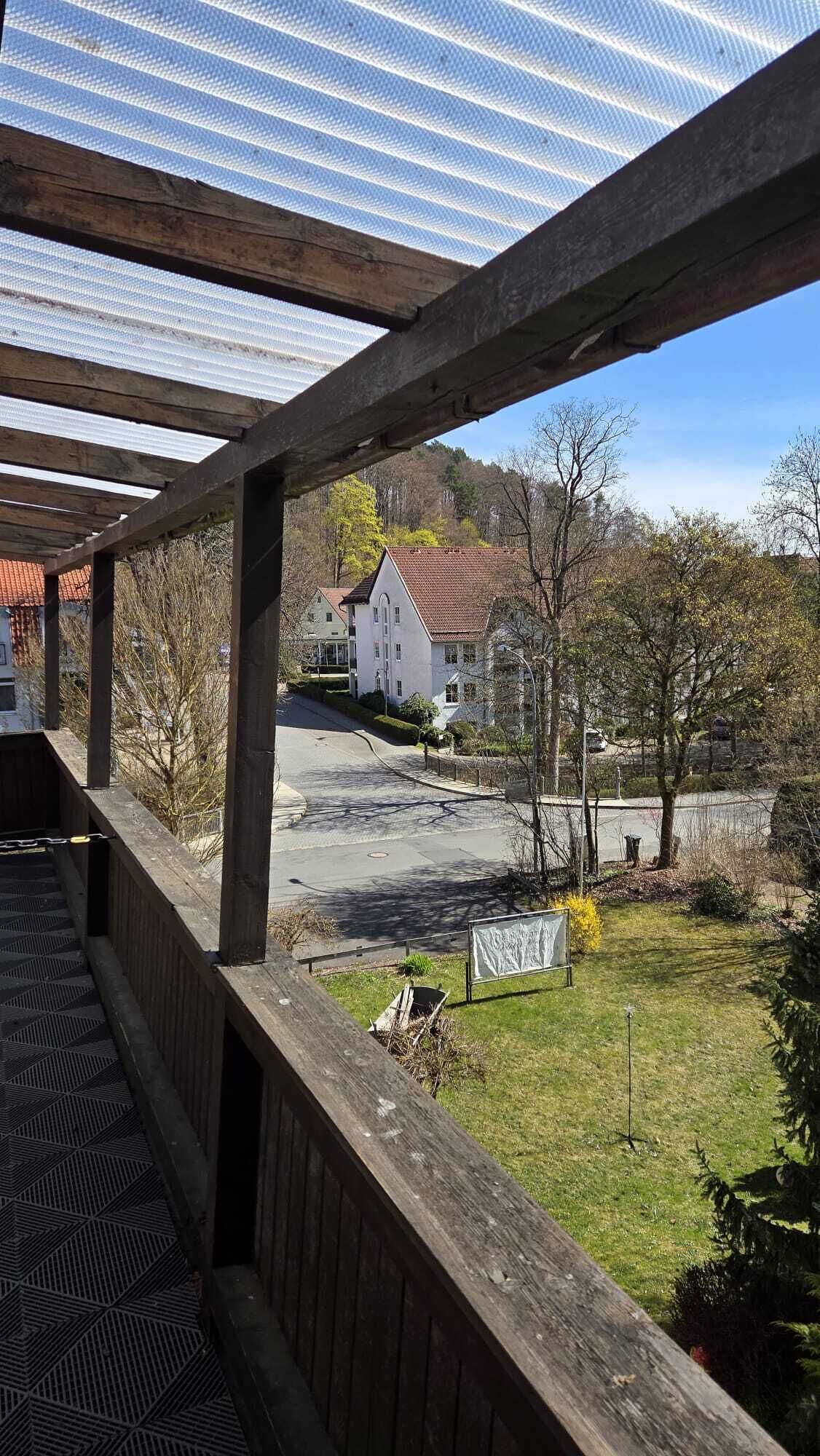 A covered outdoor area with a wooden deck, a metal roof, and a view of a residential street with houses and trees.