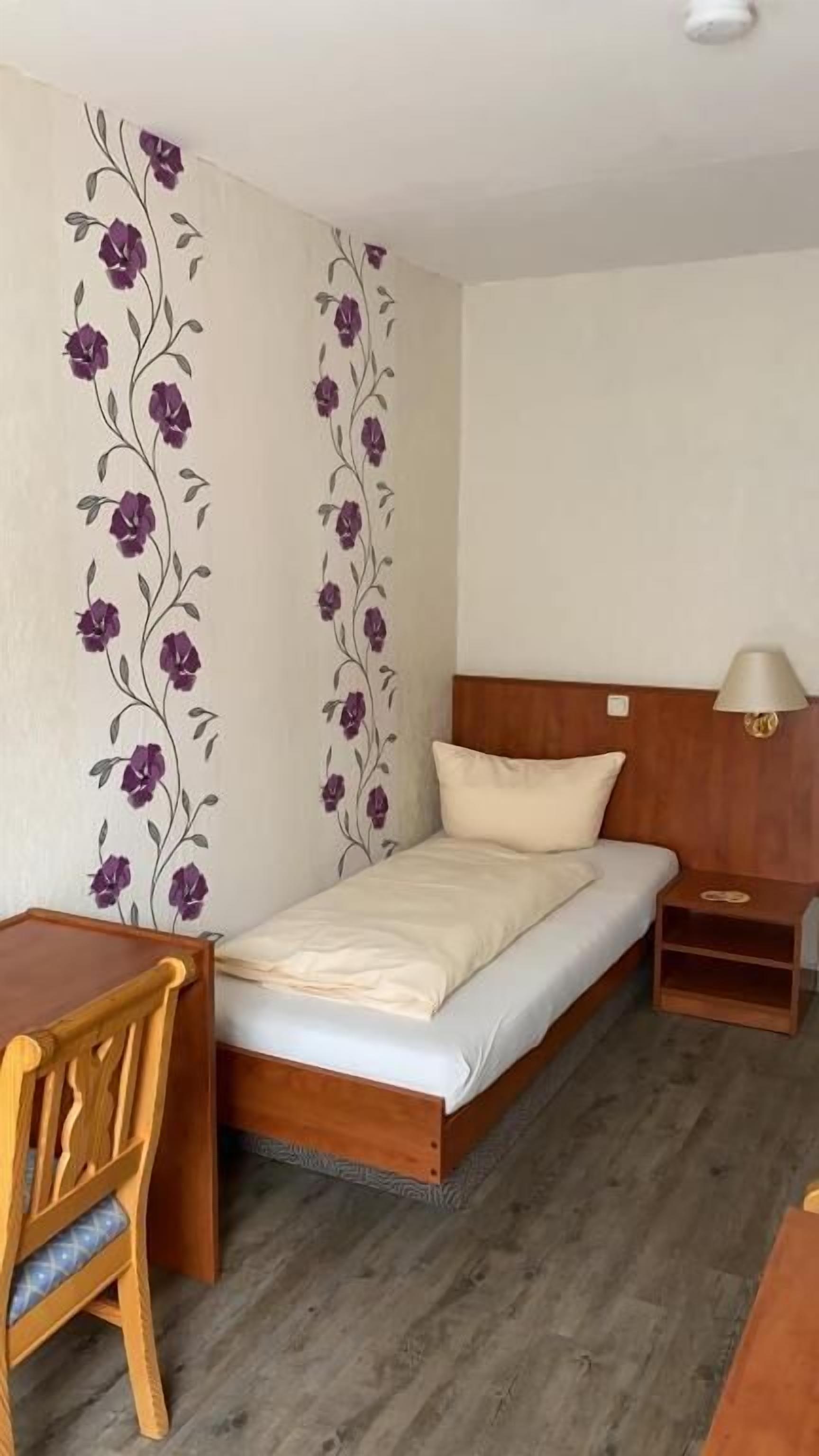 A single bed with a wooden headboard, a bedside table with a lamp, a wooden desk, and a chair.
