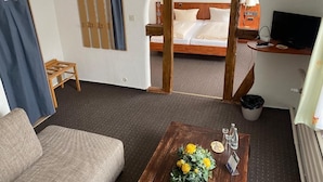 A hotel room with a bed, a desk with a computer, a coffee table with flowers, a sofa, and a view through an arched doorway.