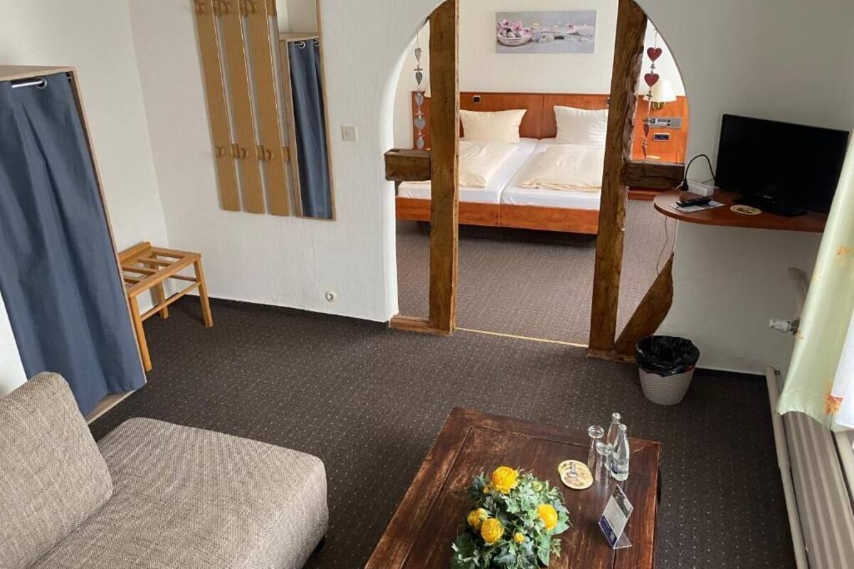 Hotelkamer met een bed, een bureau met computer, een bijzettafeltje met bloemen, een bank en uitzicht door een boogvormige deuropening.