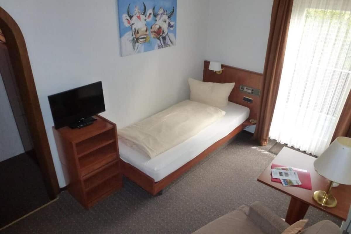 Een kleine kamer met een enkel bed, een televisie op een houten standaard, een bureau met een lamp en een schilderij van koeien aan de muur.