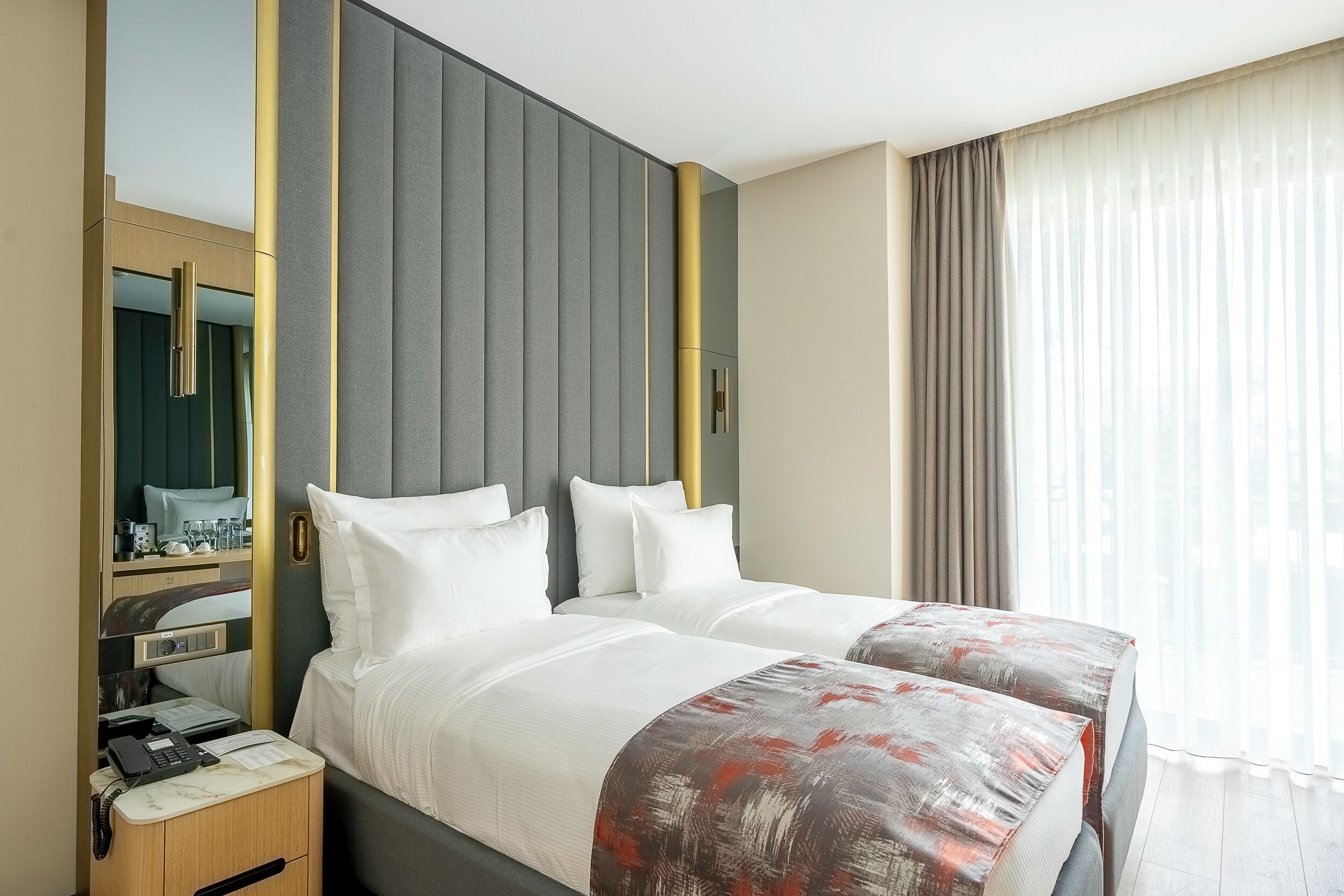 A hotel room with two beds, a desk with a phone, a mirror, and a window with curtains.