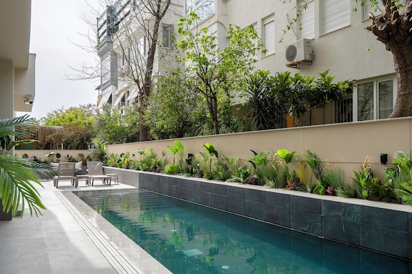 Una piscina con borde de azulejos, rodeada de vegetación y un edificio con unidades de aire acondicionado.