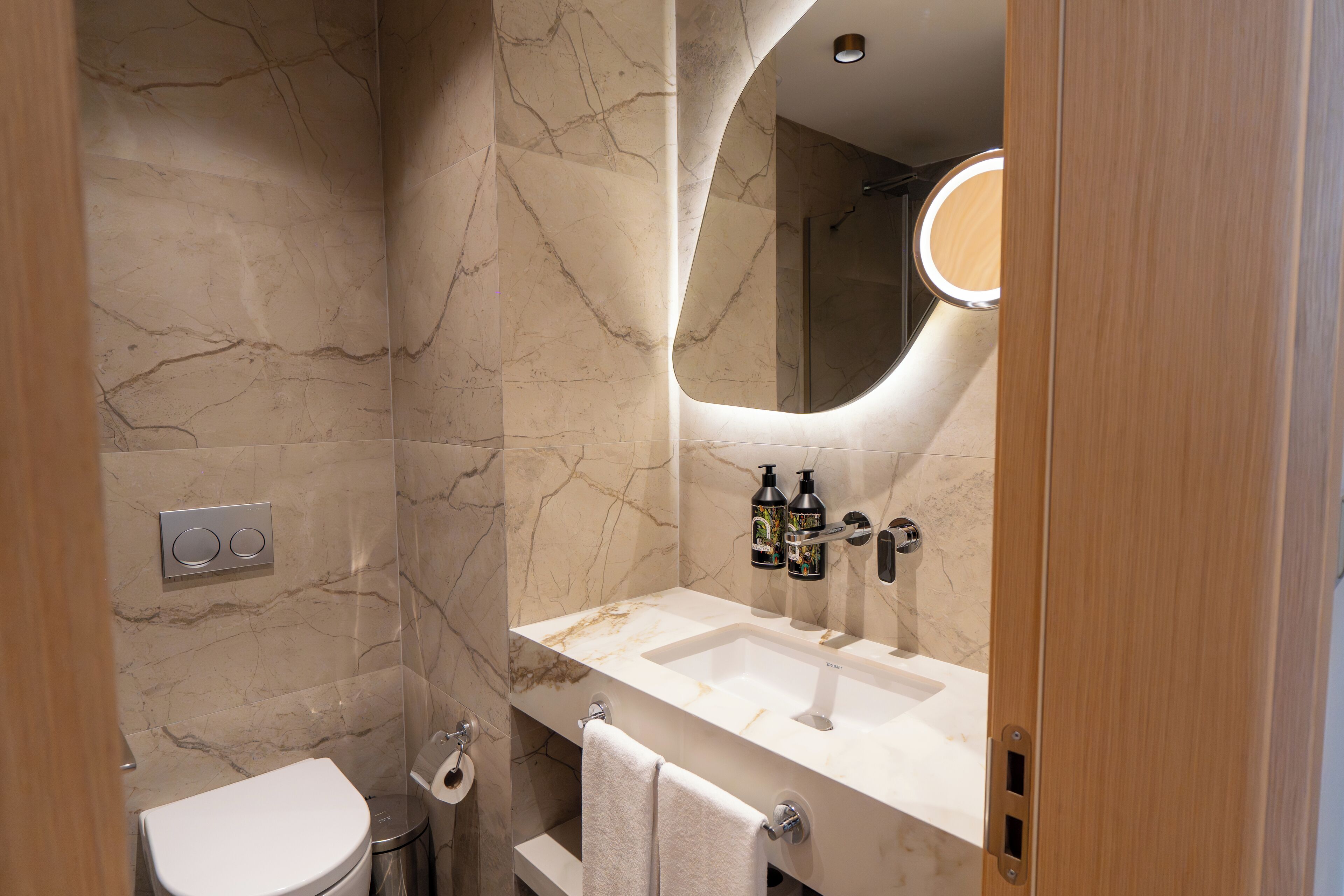 A bathroom with a marble wall, a sink with two bottles of soap, and a toilet.