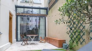 Un petit espace patio clos, avec une porte vitrée donnant sur un balcon, une petite table avec des chaises, un banc et un élément décoratif aquatique.