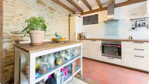 Une cuisine avec un mur en pierre, des poutres en bois, une table en bois ornée d’une plante en pot, une étagère avec des bocaux, un réfrigérateur et un tableau noir.