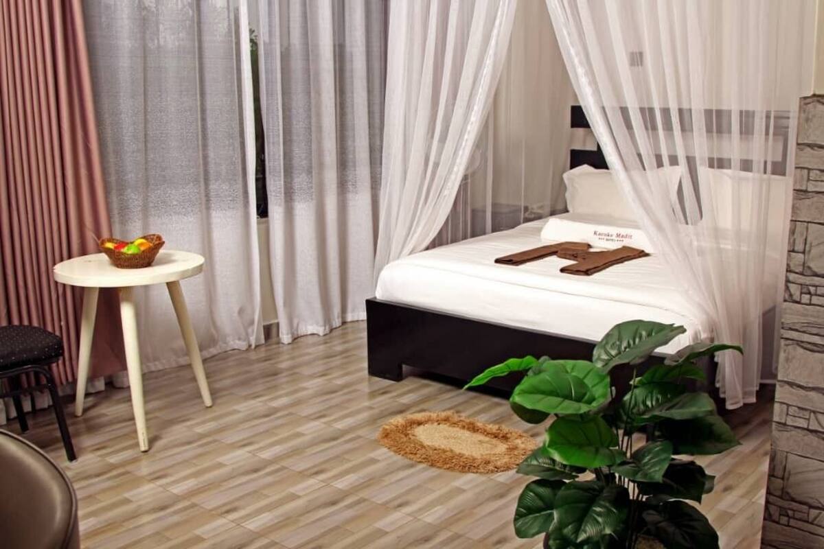 Un dormitorio ordenado con una cama, una mesita redonda pequeña, una planta en maceta y una cesta decorativa.
